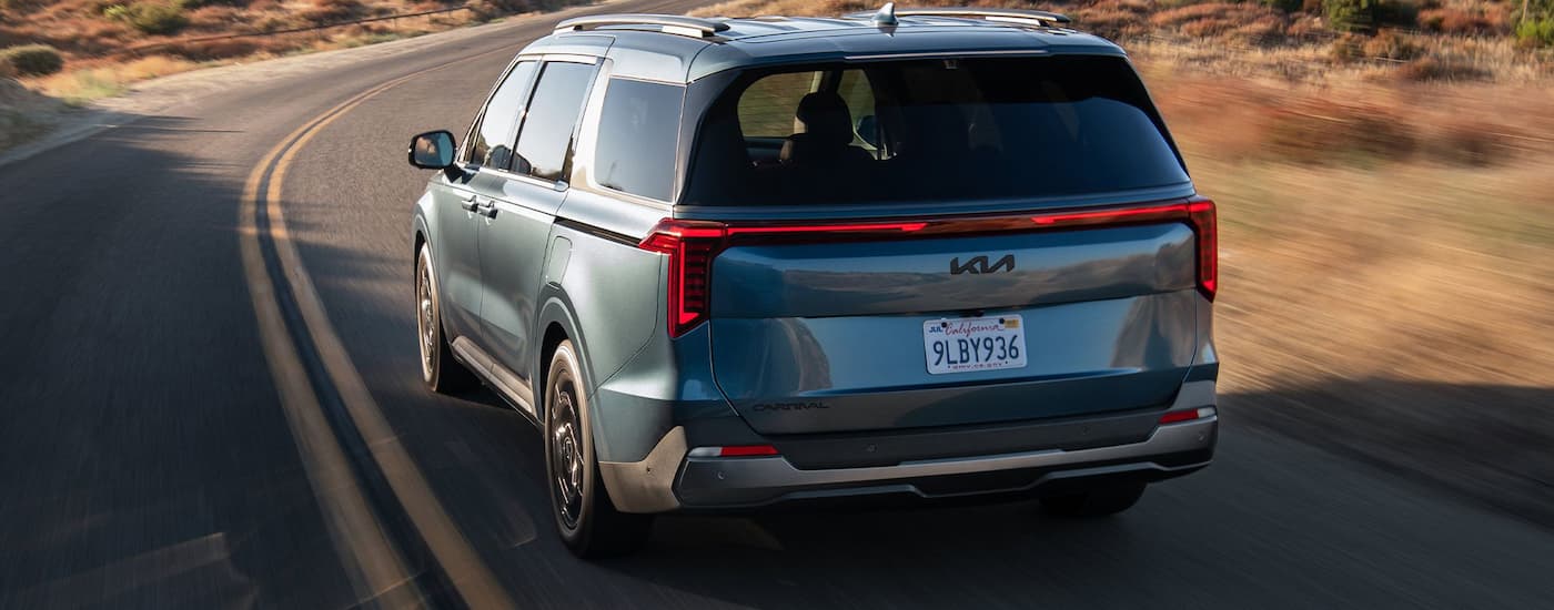 Rear of a blue 2026 Kia Carnival driving around a bend.